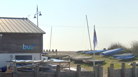 A wood cladded building bearing a blue Buzz Active logo, surrounded by stacks of paddleboards and boats and a flag, overlooks Eastbourne beach.