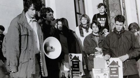 Mae'r llun du a gwyn yn dangos criw o bobl yn protestio o flaen yr hen Swyddfa Gymreig. Mae Rhun ap Iorwerth yn sefyll yno ac yn gwrando.