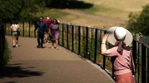 People enjoy the warm weather and sun at Greenwich Park in front of the National Maritime Museum with the Canary Wharf financial district in the distance, in London, on June 22, 2025.