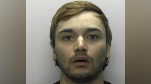 A police mugshot of Jake Ahmed. He has wavy brown hair and a vacant expression with his mouth slightly agape.