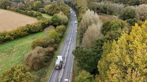 A drone view of a single carriageway stretch of road, bordered by trees and fields.