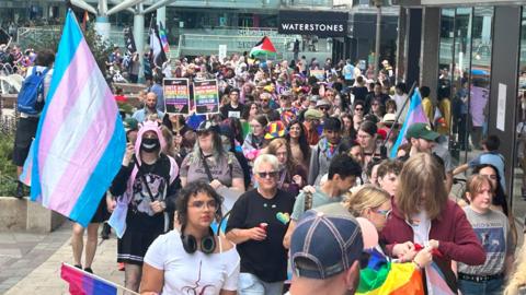 A crowd of people snakes down a city centre precinct, waving rainbow flags. Shops including Waterstones can be seen behind.