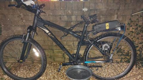 A blackened, burnt out bike that was modified with a cheap e-bike battery. There is a silver bucket of water in front of it.
