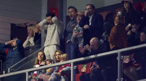Rob and Ryan at the Wrexham stadium looking disappointed as the team lost against Chelsea. There are other fans in the stands as well.