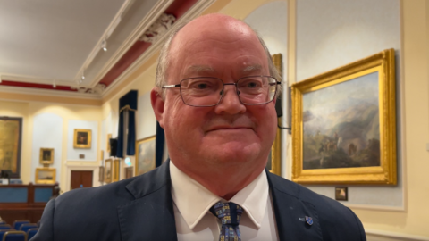 Constable of St Helier Simon Crowcroft smiles at the camera. He is wearing a blue suit with a white shirt and patterned blue tie. He is in a meeting hall which has framed paintings on the walls. He is wearing glasses.
