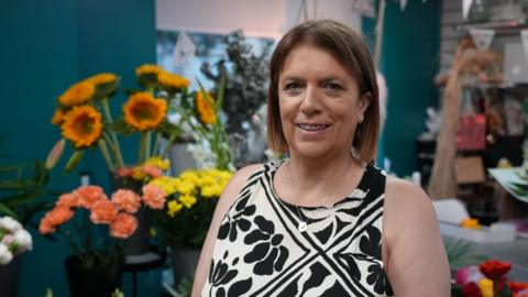 A woman with short brown hair is stood in front of flowers in her shop. She is wearing a black and white dress and looking straight into the camera.