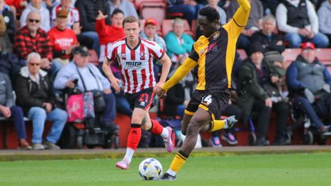 Nathan Opoku plants his left foot next to the ball, with his left arm raised as he prepares a right foot shot on the Cheltenham goal.