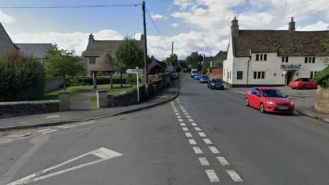 A Google street view screenshot showing the junction where the collision occurred. It is a main street going through the town, with a green space and a wooden pergola on the left, and a pub on the right. 