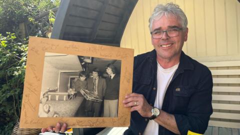 A man with gray hair wearing glasses smiles as he holds an old black and white photograph on a cardboard mount with signatures around it. The photo shows him as a child with Graham Taylor, a dark-haired man wearing a blazer and tie, and a taller man with a dark mullet wearing a tie and jumper.