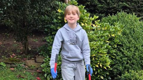 Young boy standing in a garden, smiling at the camera while holding two litter pickers, wearing a grey hoodie, blue gloves and casual trousers, with green bushes and trees behind him