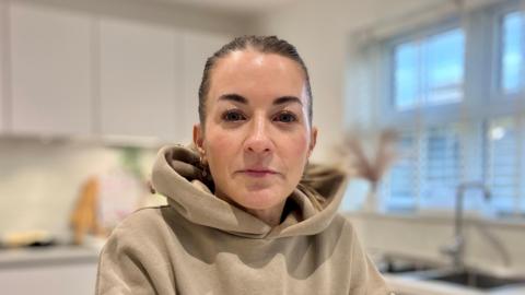 A person wearing an oversized beige hoodie seated at a white countertop in a modern kitchen. The kitchen features white cabinets, a sink under a large window with blinds.