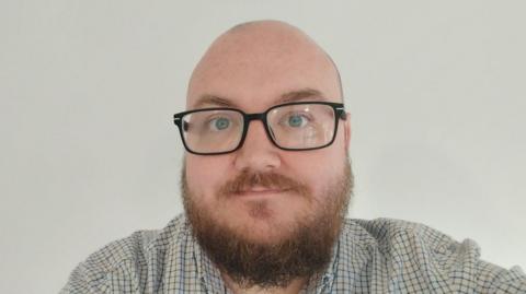 A selfie of a man wearing glasses and a shirt while standing in front of a white wall. 