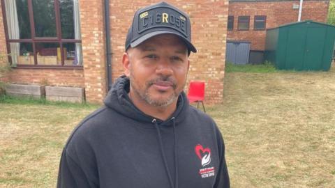 A man in a black hoodie and black cap looks at the camera. 
