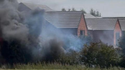 Black smoke in front of a new housing development. The smoke is spreading across a nearby field. 