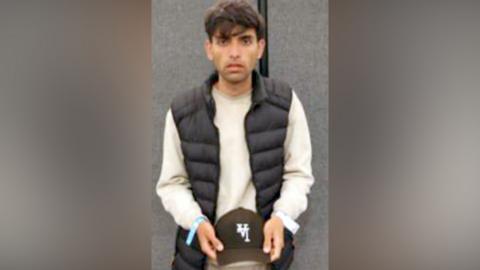 A young man stands in front of a camera. He is holding a cap in his hands.