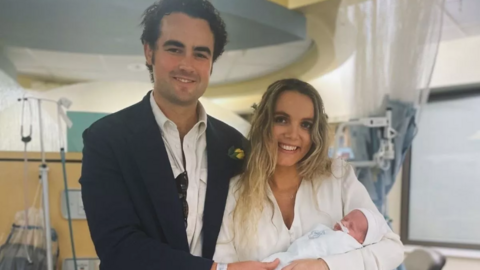 Archie and Issy Sylvester stand in a hospital room smiling at the camera. The mother has her young baby in her arms. He is wrapped in a blanket with a white hat on and has some tubes stuck to his face. The couple are smartly dressed, with the man in a white shirt and suit jacket while the woman is dressed in white.