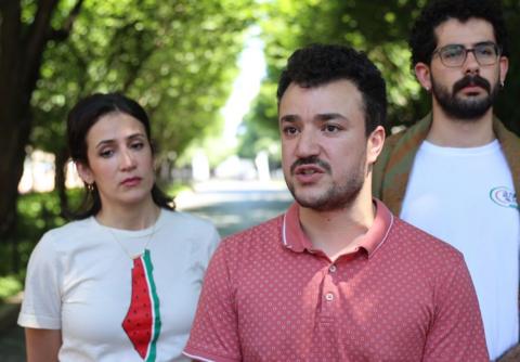 Mahmoud Khalil wears a pink shirt outdoors and is flanked by two people in white shirts. 