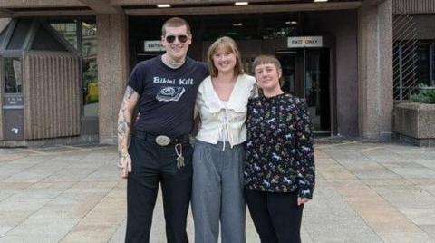 A man stands next to two woman outside Liverpool Crown Court. They have their arms around each other and are smiling. 