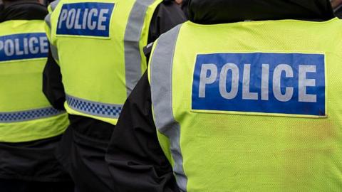 Stock photo of a police man facing away form the camera. He is wearing a black jacket with a yellow high vis jacket that reads POLICE on the back.