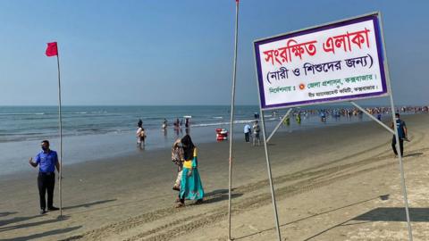 People walk past a signboard reading "Reserved area for women and children as instructed by the district commissioner" along a beach in Cox's Bazar after authorities in Bangladesh's main resort district scrapped a dedicated beach section for women and children after a social media outcry over gender segregation, officials said on December 30, 2021.