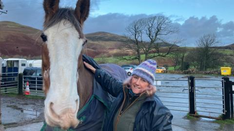 Cumbrian Heavy Horses riding centre owner Annie Rose standing in the yard of the riding centre with one of her horses. The horse is a large, heavy horse breed and is brown with a white face. Rose is resting her hand on the horse's shoulder.