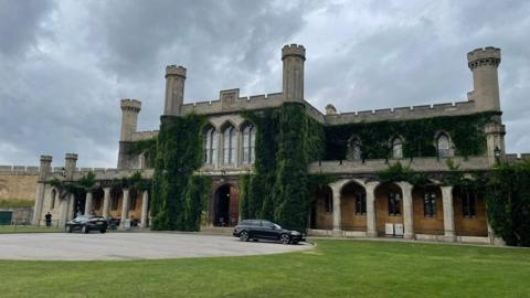 An image of the exterior of Lincoln Crown Court, a large historic building with columns, turrets and an ivy covering. There is a lawn outside with two black cars parked in front.