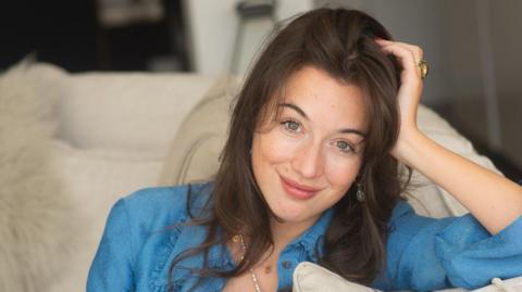 A woman with brown hair smiling wearing a blue blouse sat on a white sofa. 