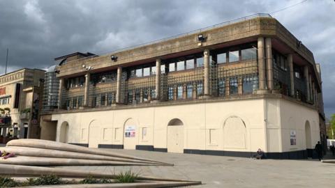 A large three-storey building with columns. Its first floor is boarded up. The building sits in front of a paved square. 