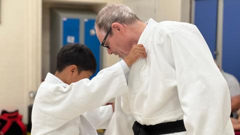 Tony is pointing at a child's feet and giving instructions to the youngster. Tony has grey hair and is wearing glasses. He has a black belt around his white judo outfit. The child has black hair and is also wearing a white judo strip