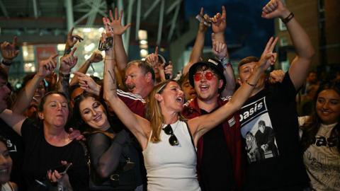 Grŵp o bobl yn sefyll yn agos at ei gilydd mewn ar ol cyngerdd Oasis yng Nghaerdydd, gyda goleuadau nos yn y cefndir. Mae sawl person yn codi eu dwylo yn yr awyr, rhai yn gwneud arwyddion heddwch ac yn dal ffonau symudol. Dillad fel crysau-t gyda logo Oasis. Yn y cefndir, gellir gweld strwythur stadiwm y Principality a rhai adeiladau brics.