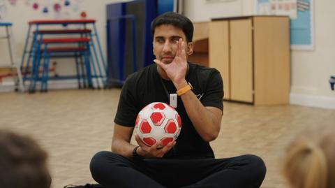 An instructor for blind football holding a ball in a school sports hall with children watching.