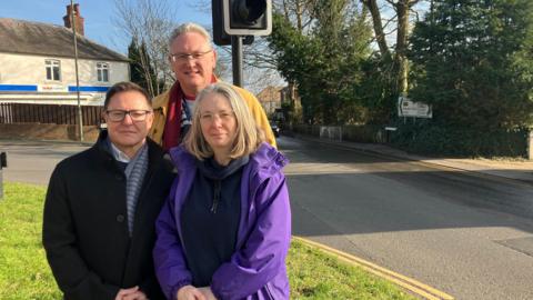 Councillor Catherine Powell, with residents Elliot Russo and Simon Cross standing by the traffic lights on Upper Hale Road. In the background is a branch of Tescos and signs directing cars where to go.