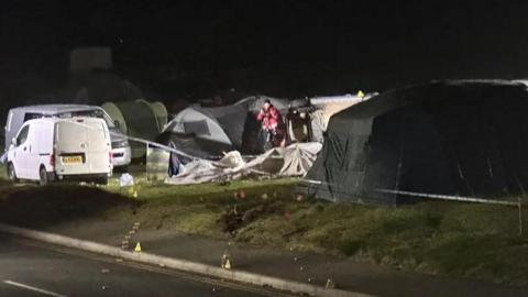 A group of tents can be seen in a field with one central tent seen crushed. They are all surrounded by a police cordon with markers on the road recreating the crash.