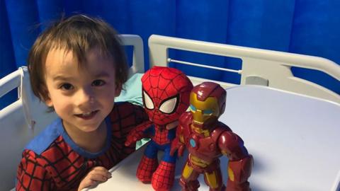 Theo Sebastian-Jenkin, then four-years old, sitting up on a hospital bed wearing a spider man costume while staring straight at the camera and playing with super hero toys.