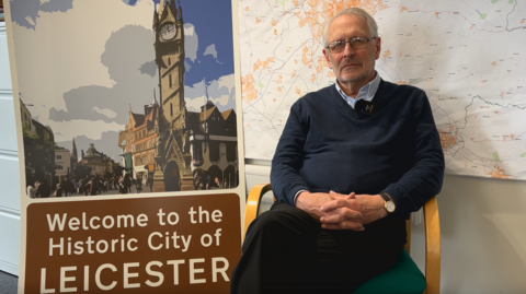 Sir Peter Soulsby sits in front of a map of Leicestershire with a sign saying "Welcome to the historic city of Leicester" at his side