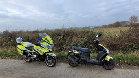 A police motorcycle and a stolen moped scooter. Hedges and a field are in the background.