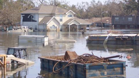 A flooded street in New Orleans with damaged houses, debris and an overturned vehicle