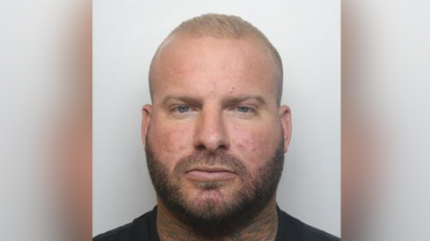 Mugshot of Alex Male in a black t-shirt. He has short blond hair, a light brown beard and a neck tattoo