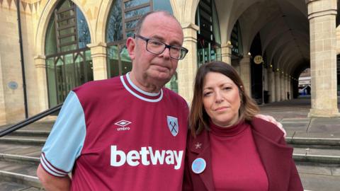 A man in a claret and blue football shirt stands close to a woman in a maroon blazer and red jumper in front of an old building.