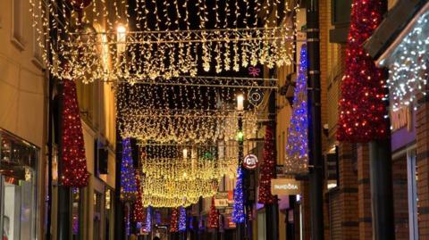 Strings of gold lights hang down along the length of Strand Street, with blue and red illuminated tree-shaped decorations on either side.