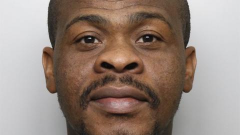 A mugshot of Abass, who is wearing a grey sweatshirt and has a bald head. The background is plain and light-coloured, 