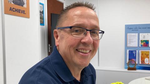 A middle aged man wearing glasses smiles toward the camera , he is wearing a blue polo shirt.