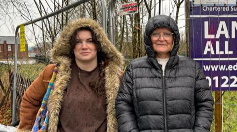 Bethany Wood, with dark hair, wearing a fur-lined hooded coat, standing next to Helen Fisher, who is wearing glasses and has a black puffer coat on with the hood up. Behind them is a metal fence with a sign that reads land for sale, sold. There is overgrown grass and bushes behind the fence. It is a wet day.