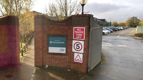 A brick entrance with iron gates, with a car park seen in the distance The wall has an NHS-branded sign reading: "Welcome to Cheswold Park." Road signs warn of speed bumps, a five miles an hour speed limited an instruction for all vehicles to turn left. 