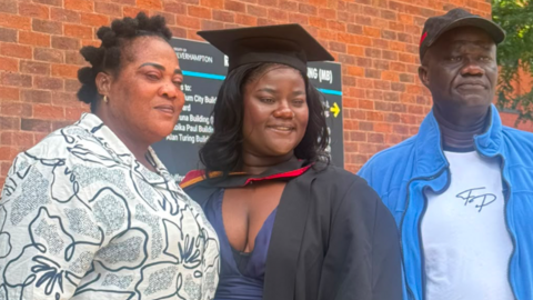 A woman in the middle is wearing a black graduation gown. Another woman on the left is wearing a dark and white top, while a man on the right is in a dark baseball cap and blue and white clothing.