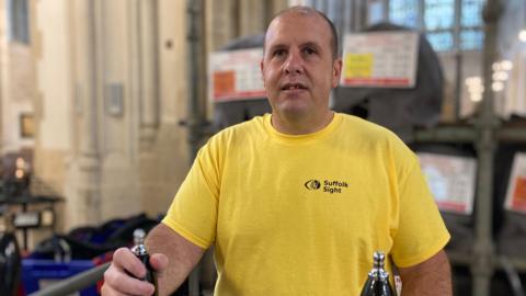 Kevin Ramsey stands behind a bar inside St Edmundsbury Cathedral. One of his hands rests on a beer tap lever on the bar. He has a shaved head and wears a yellow T-shirt with the words and logo of Suffolk Sight on the right hand side of his chest. 