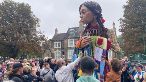 The puppet is surrounded by people taking photos as she makes her way through the park. She is draped in a patchwork blanket.