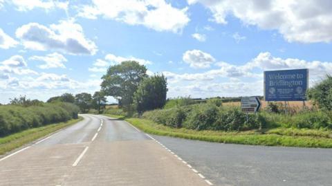 A country road flanked by green bushes and trees. There is an exit lane on the right with a big blue sign on the grass verge reading "Welcome to Bridlington". Next to that is a white road sign listing Grindale, Burton Fleming and Malton, with a black arrow pointing right. The sky is blue with scattered white and grey clouds.