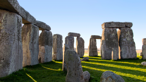 The inner-stone circle at Stonehenge with shadows cast on the grass and sunlight coming through the gaps in the stones with blue sky in the background.