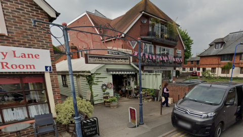 A florists shop in a light green timber building. There are rows of flowers outside. There is a metal sign saying "to the shops" outside that is over a pathway down the side of the florists. There is a black car parked outside the shop. 